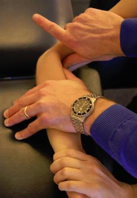The clinician provides a lateral to medial glide while the patient pushes their own wrist into further