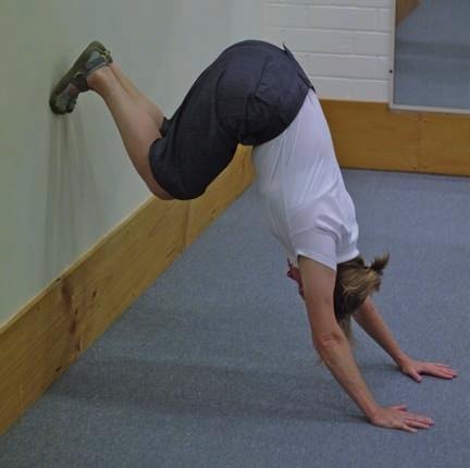 Practise handstands chest-to-wall, not back-to-wall; in the latter, it is very difficult to produce the correct line through the body. Have your hands shoulder-width apart on the floor.